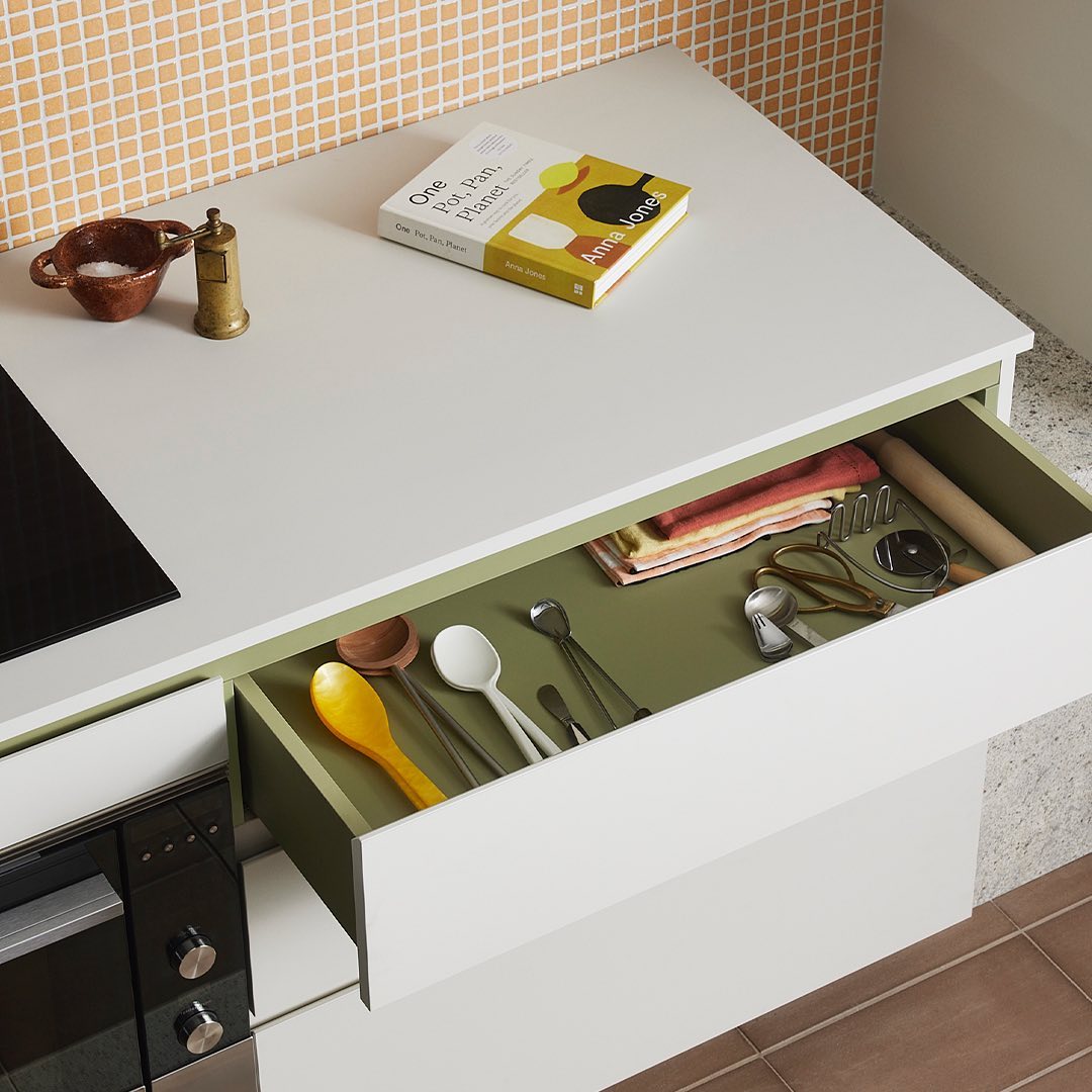 Kitchen interior with white counter, open drawer with utensils, cookbook, salt, and pepper grinder; light-orange square tile backsplash.