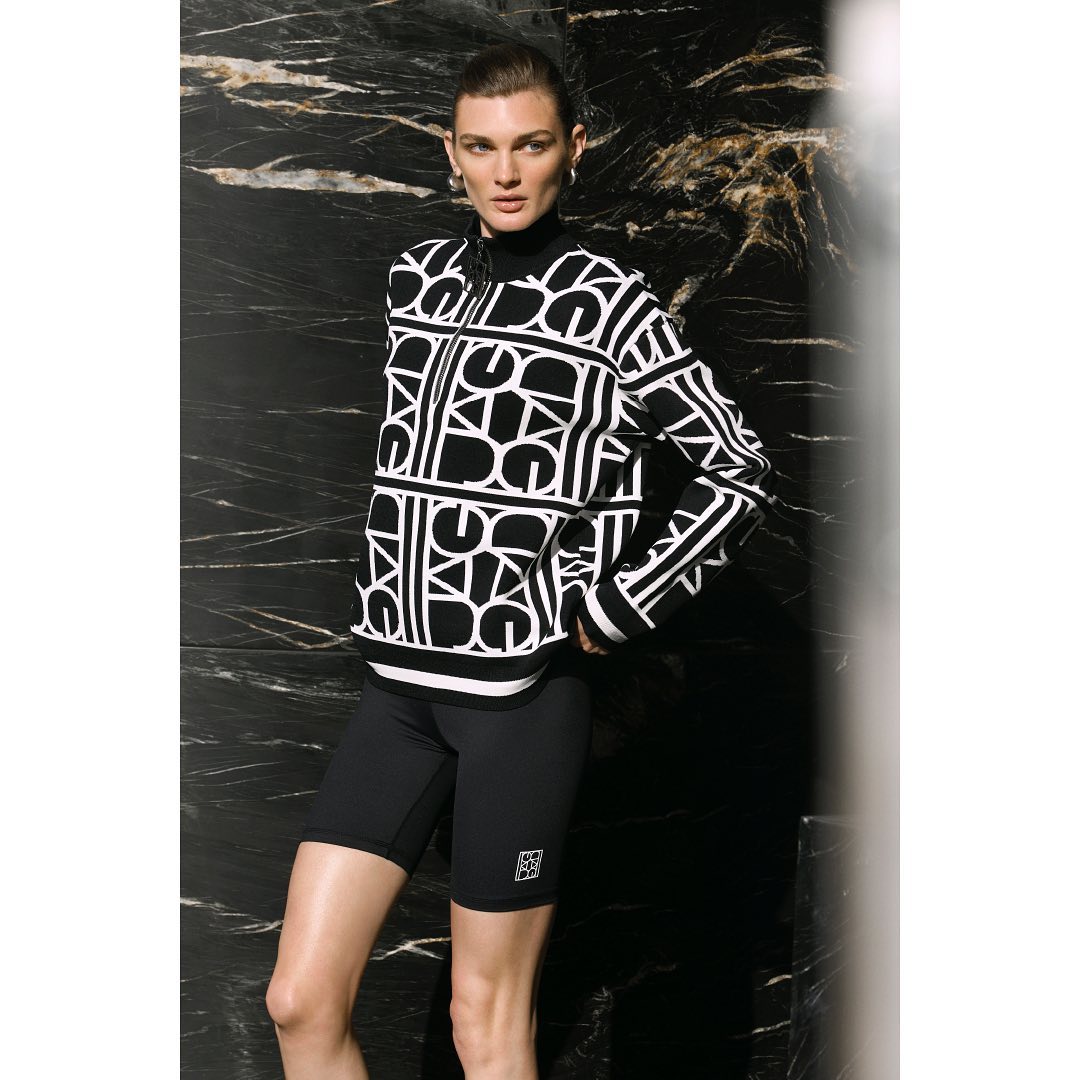 A fair-skinned woman in black bike shorts and a black and white geometric-patterned zip sweater poses against a dark marble wall.