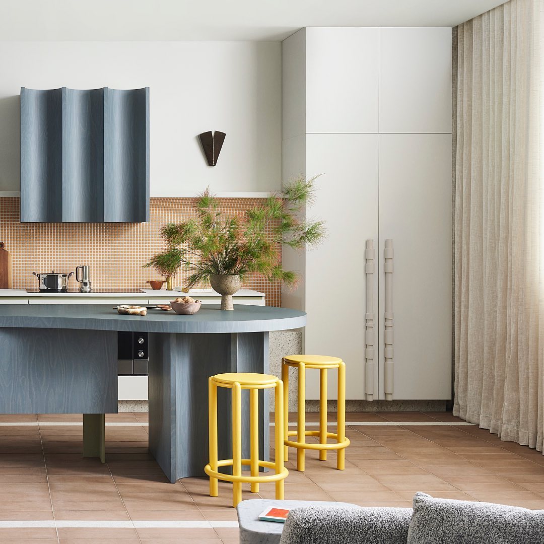 Contemporary kitchen interior featuring a blue island with stools, light peach tiled backsplash, and neutral color palette; indoor daylight.