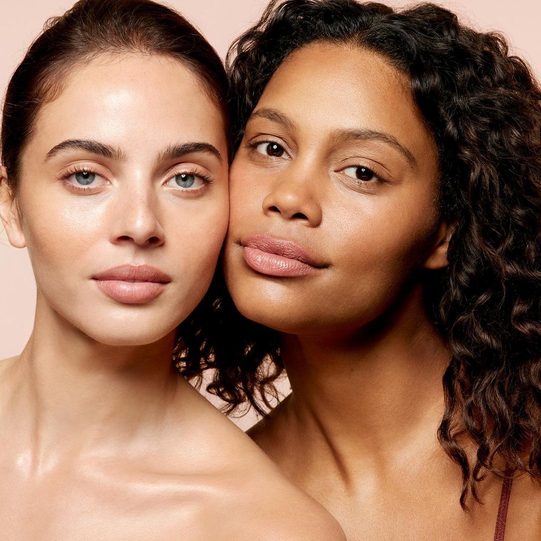 Close-up of two women with diverse skin tones posing closely together, showcasing smooth skin, natural makeup, and a soft, neutral background.