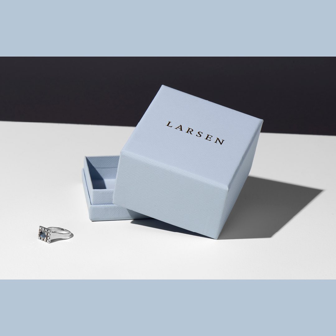 Jewelry product shot featuring a silver ring with a gemstone next to a light blue Larsen branded box on a white surface with a dark background.