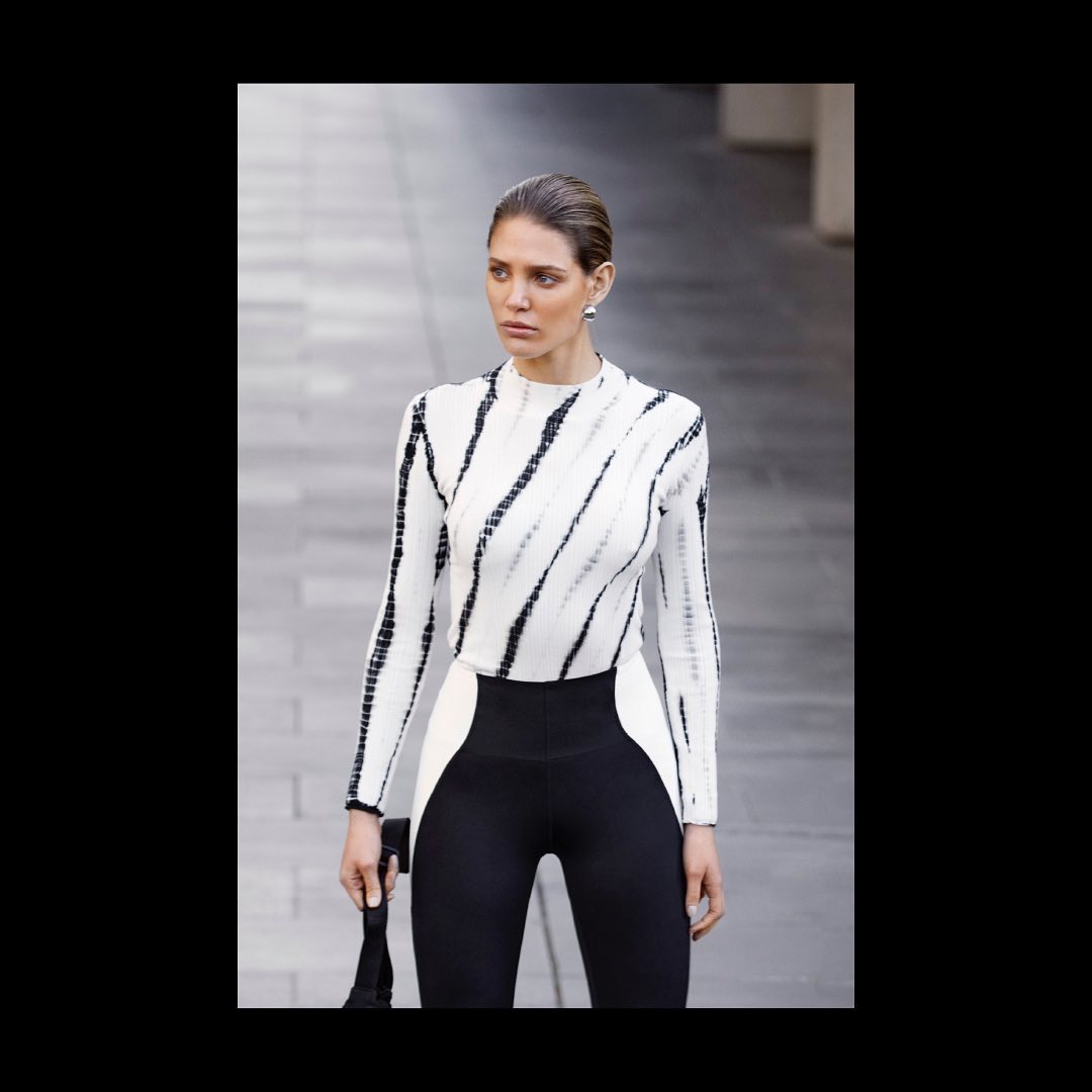 Stylish woman in black and white long-sleeve top and leggings poses on city street with blurred background of grey architecture.
