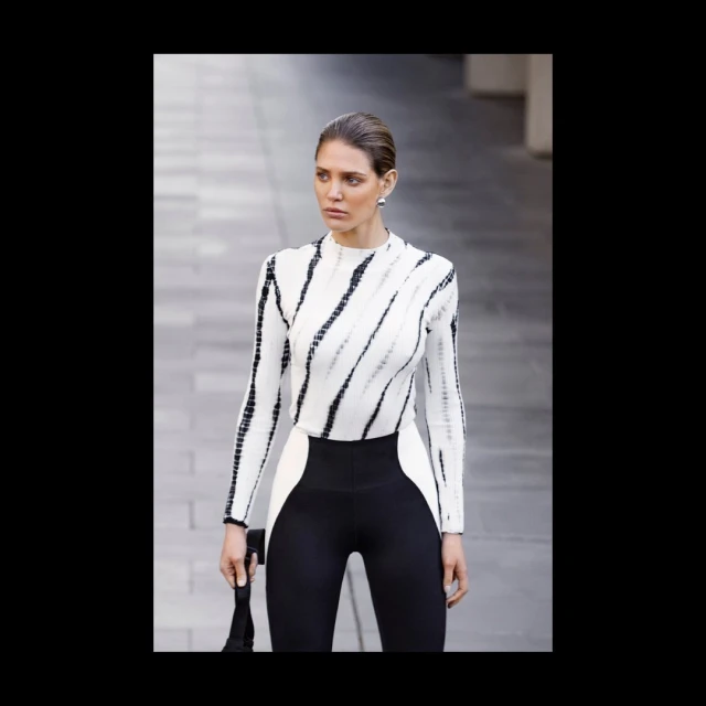 Stylish woman in black and white long-sleeve top and leggings poses on city street with blurred background of grey architecture.