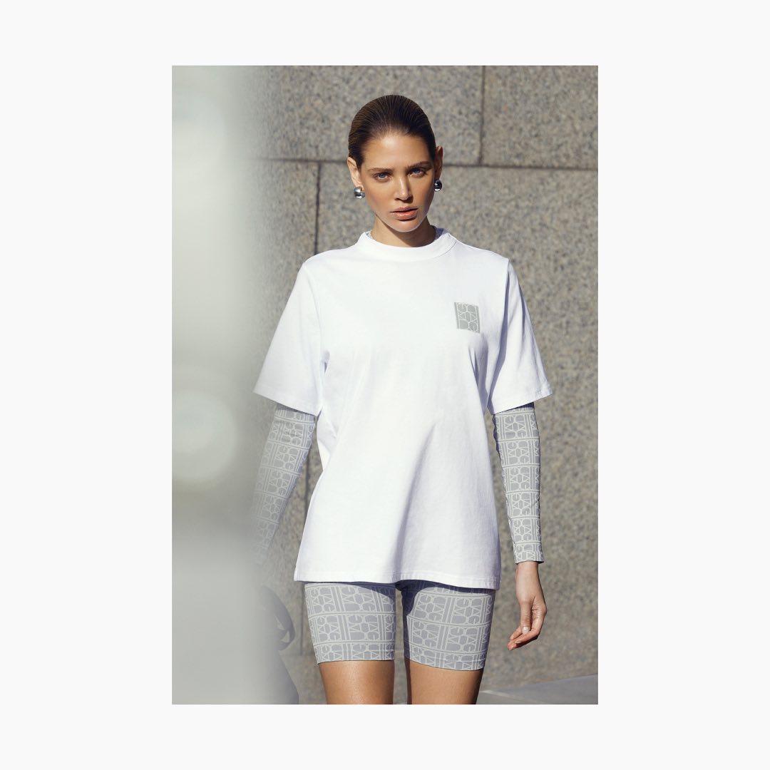 A young woman stands in front of a concrete wall, wearing a white T-shirt and gray patterned shorts. She has her hair pulled back.