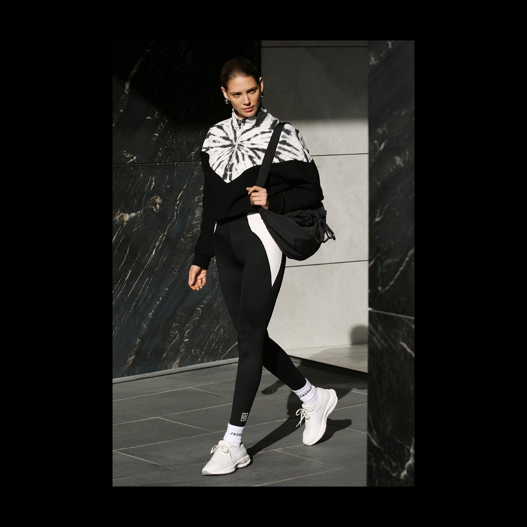 A woman in a black-and-white tie-dye sweatshirt and black leggings walks on a city sidewalk, carrying a black duffel bag over her shoulder.
