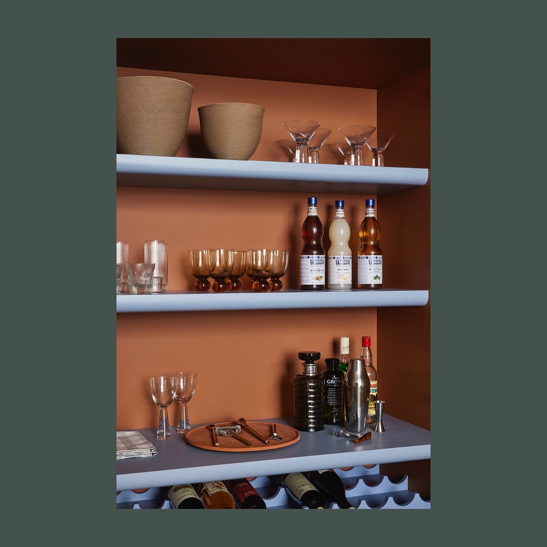 Interior view of a built-in bar shelf with liquor bottles, glassware, and bar tools, against a backdrop of terracotta-colored wall.