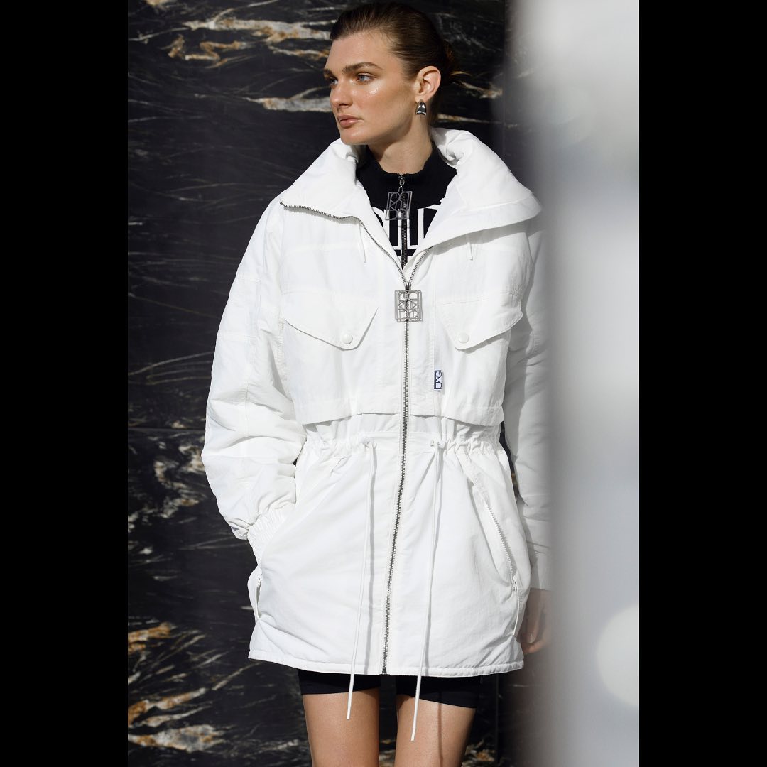 A woman in a white puffer jacket is centered against a dark, textured background. She wears earrings and a necklace.
