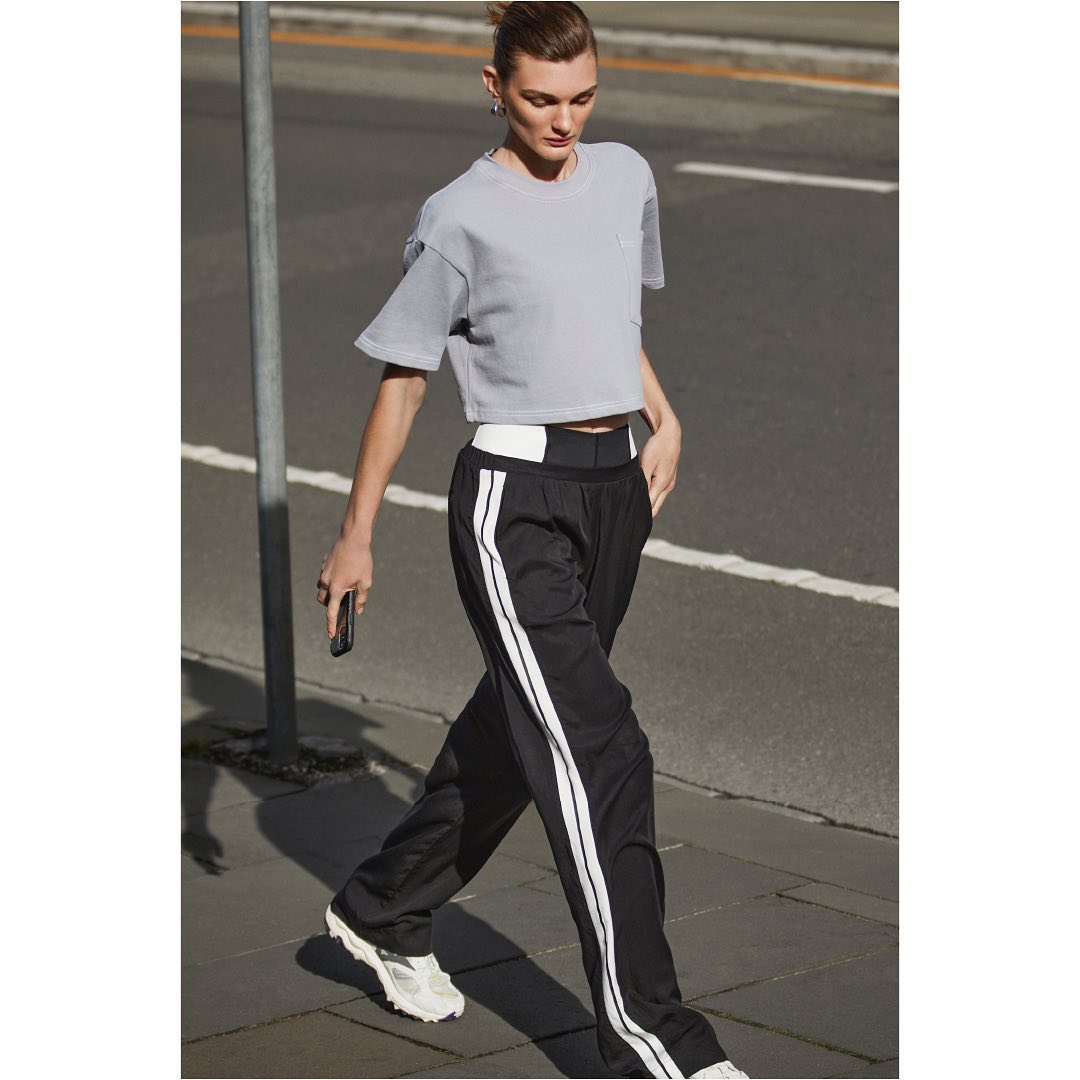 Woman in a grey t-shirt and black athletic pants walks on a city street holding a phone, with blurred background details and natural lighting.