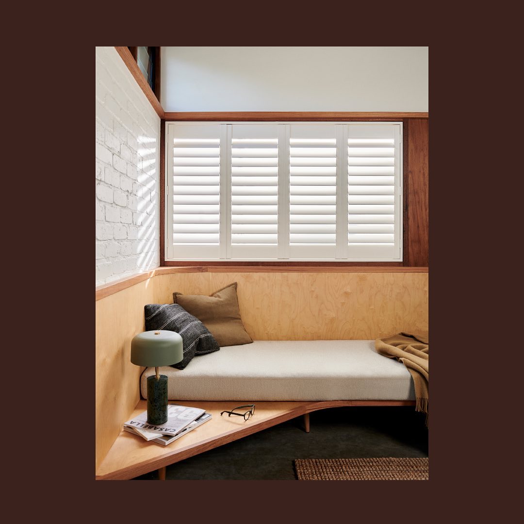 Corner daybed nook with natural light from window with plantation shutters, throw pillows, books, and lamp. Cozy indoor space.