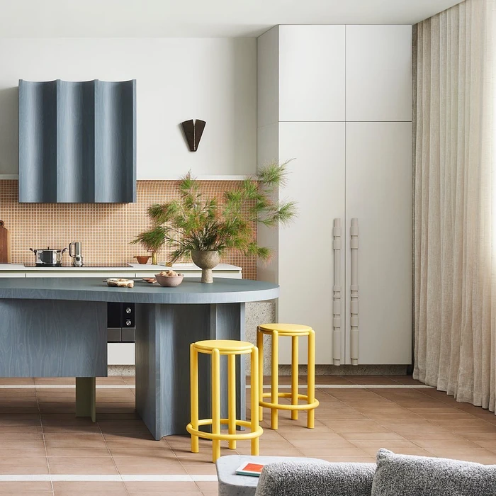 Modern kitchen with a curved slate blue island, two yellow stools, and tall white cabinetry by Ortolan in Melbourne.