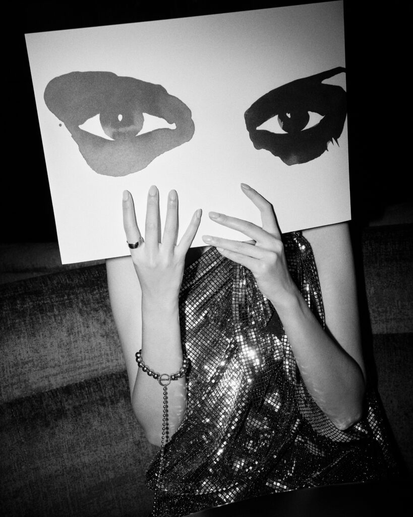 Person in a black and white photo holding an art board with painted eyes, wearing a shiny top with rings and a bracelet.
