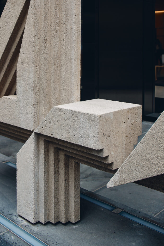 Close-up view of a large, beige geometric outdoor sculpture made of stone or concrete, set against a dark building facade.