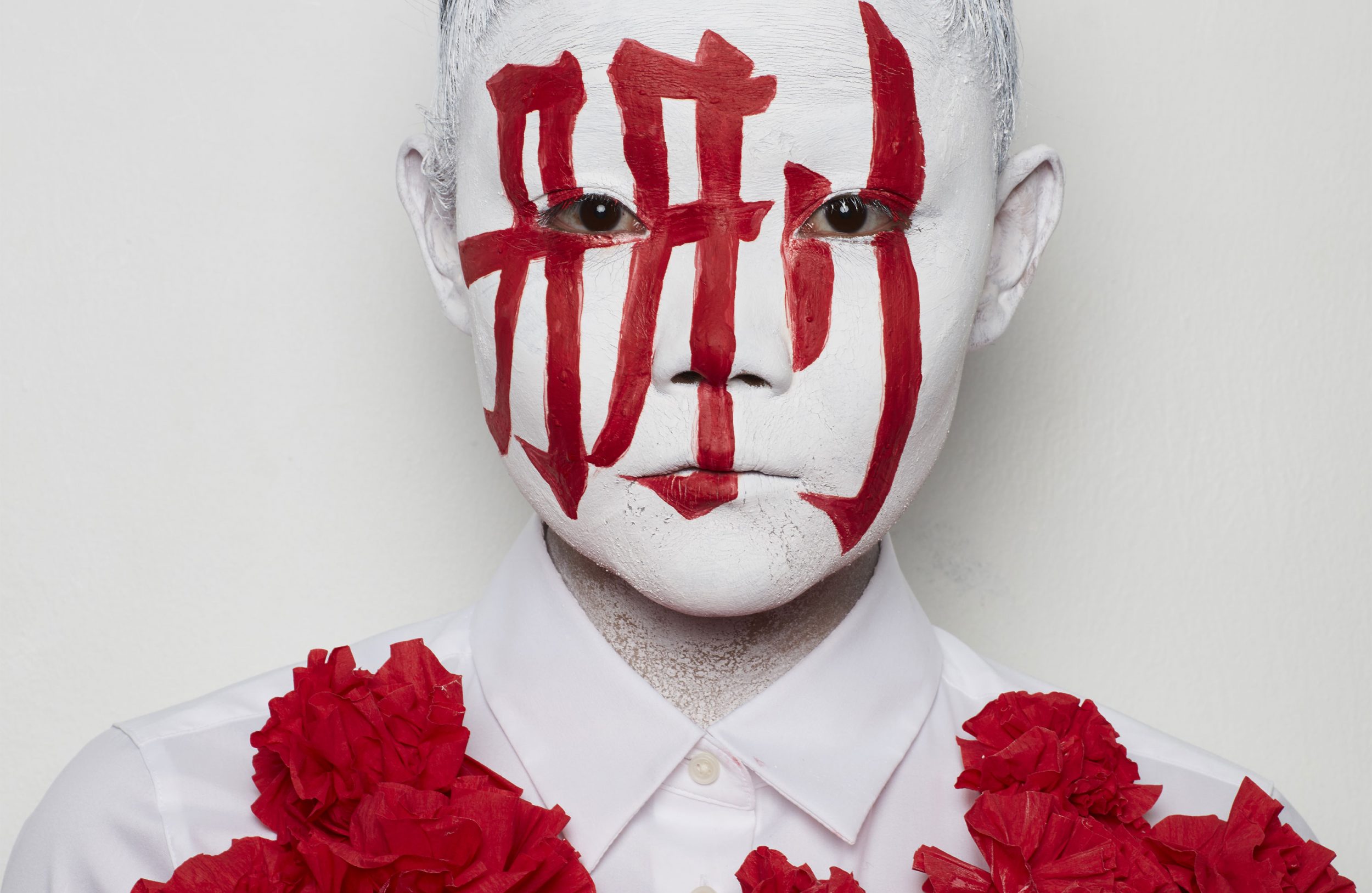 Artistic portrait of a person with white face paint and red painted Kanji characters, accentuated by red floral embellishments.