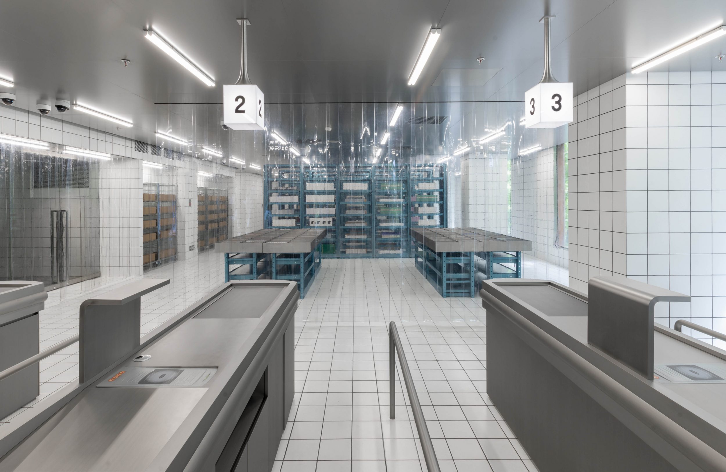 Empty commercial kitchen area featuring tiled walls, metal tables, shelving, and hanging number signs inside of a food production facility.