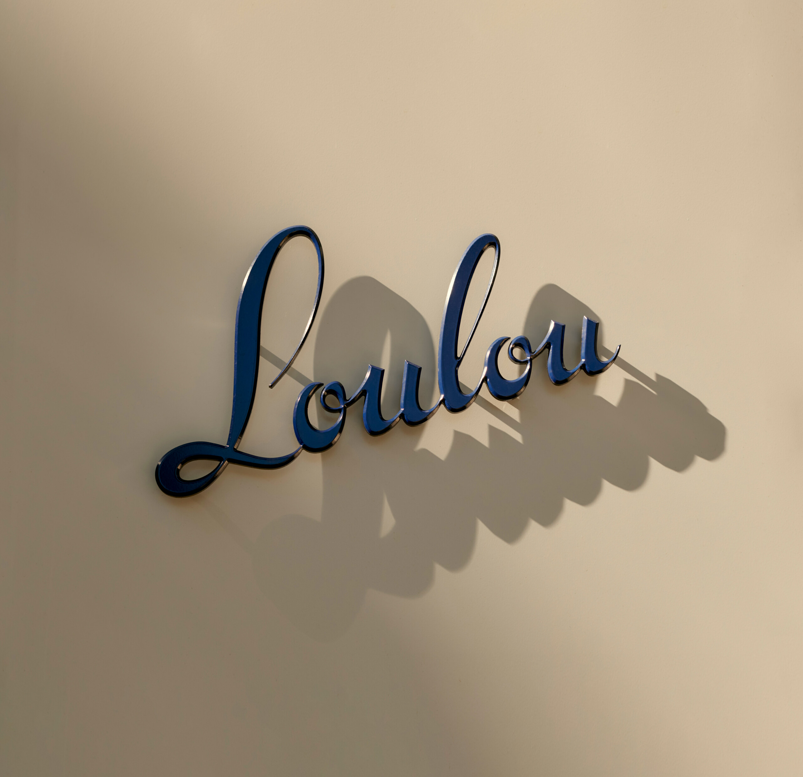 Blue cursive script 'Loulou' on a light surface, casting a shadow; minimalist composition with a focus on typography and design.