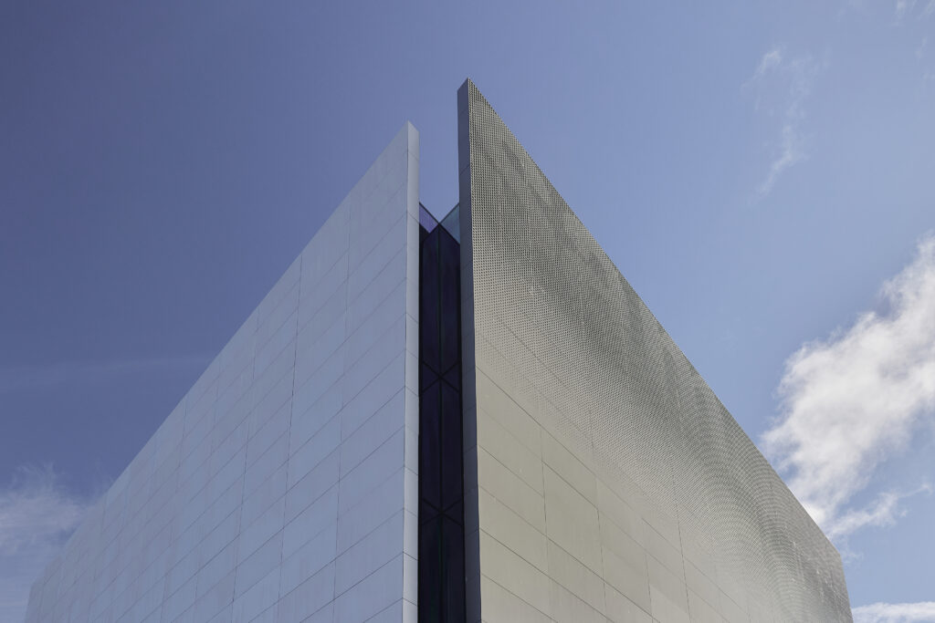 Exterior view looking up at a modern building featuring clean lines, geometric shapes, and blue sky with light clouds in the background.