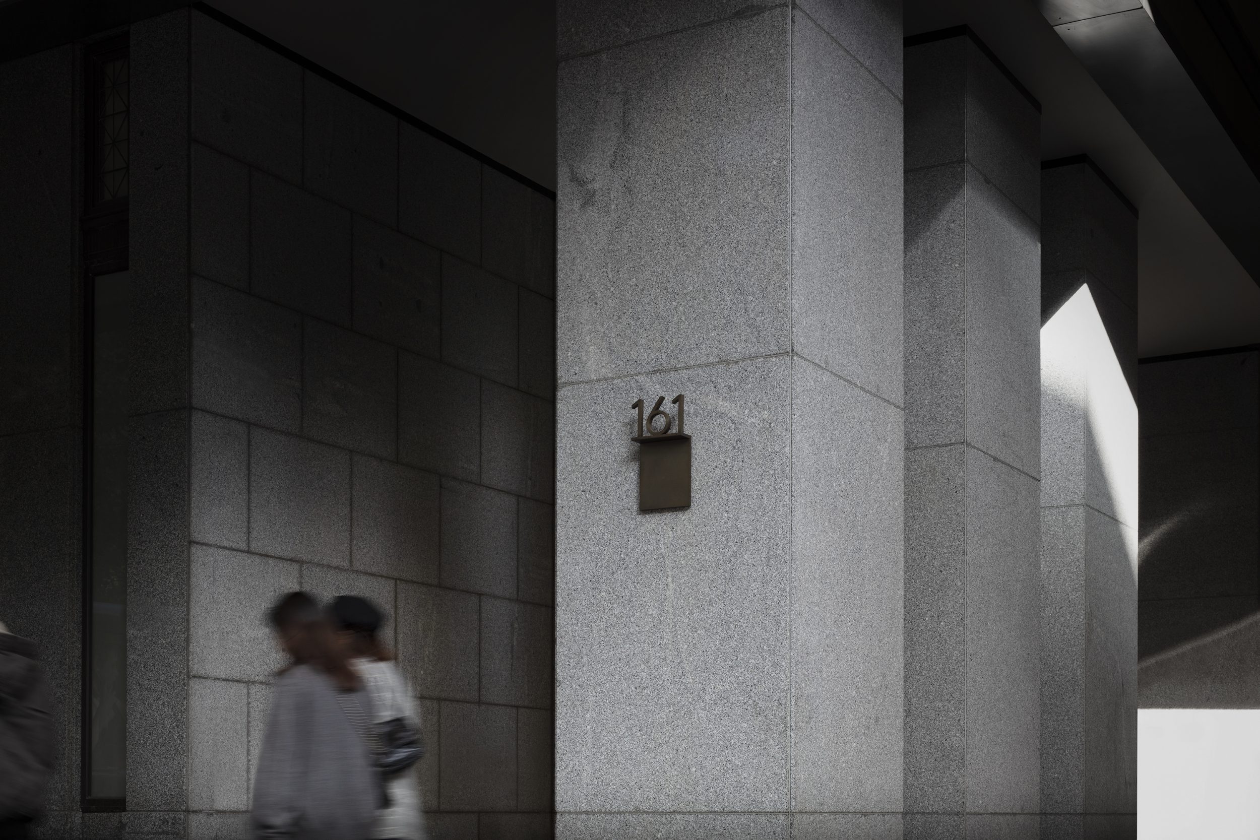 Blurred figures walk past concrete building columns marked with '161', creating an urban street scene with geometric shadows and monochrome tones.