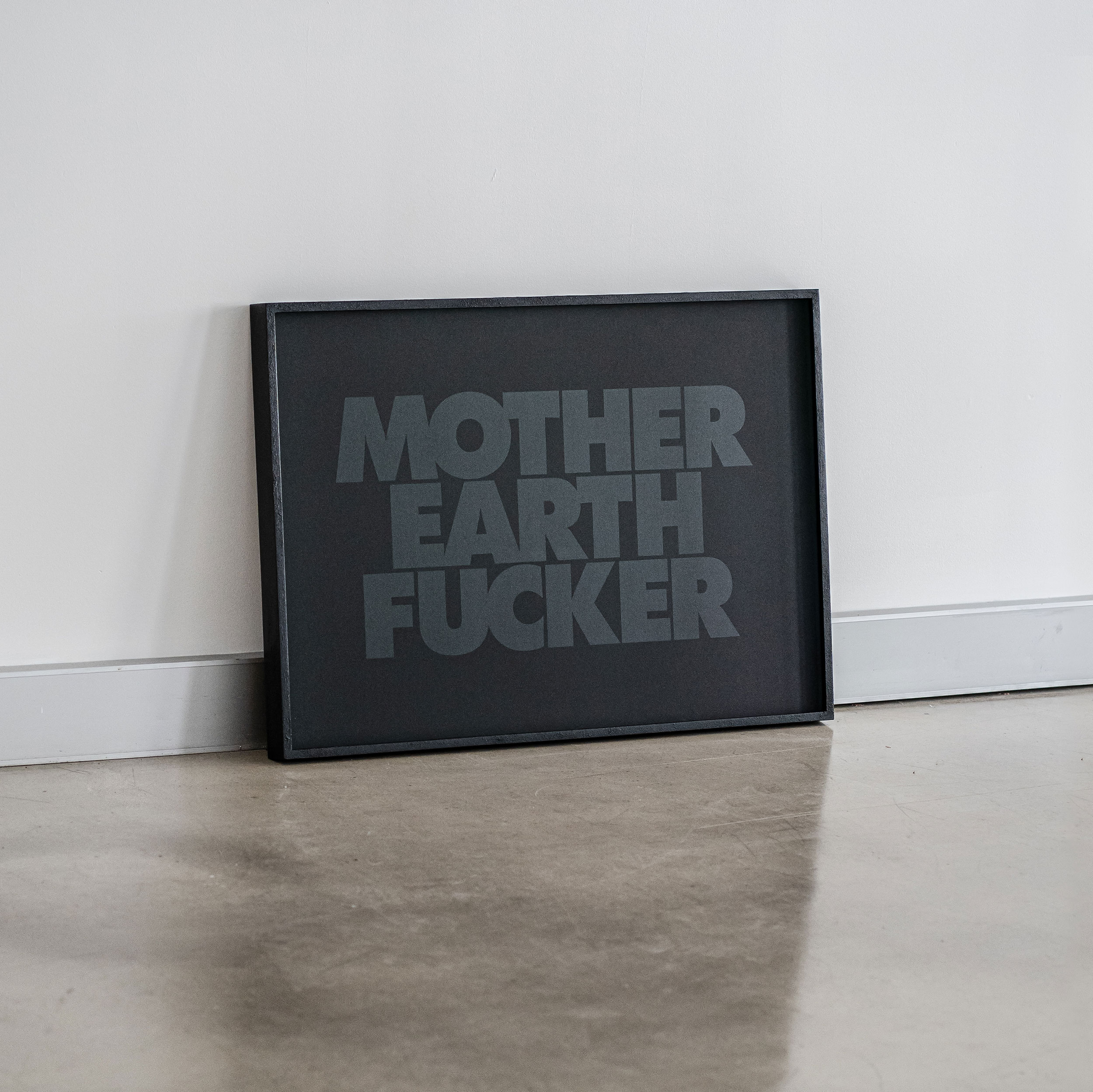 Framed black and grey print with the text 'Mother Earth Fucker' leaning against a white wall on a grey concrete floor.