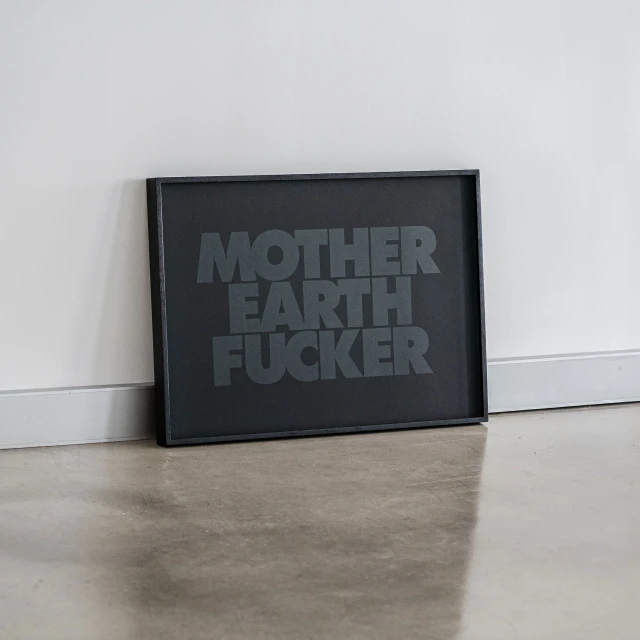 Framed black and grey print with the text 'Mother Earth Fucker' leaning against a white wall on a grey concrete floor.