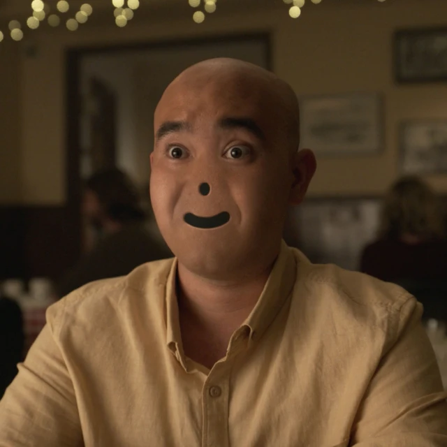 Close-up of an adult male with a shaved head and a simulated smiley face, wearing a beige button-down shirt in a dimly lit indoor setting.