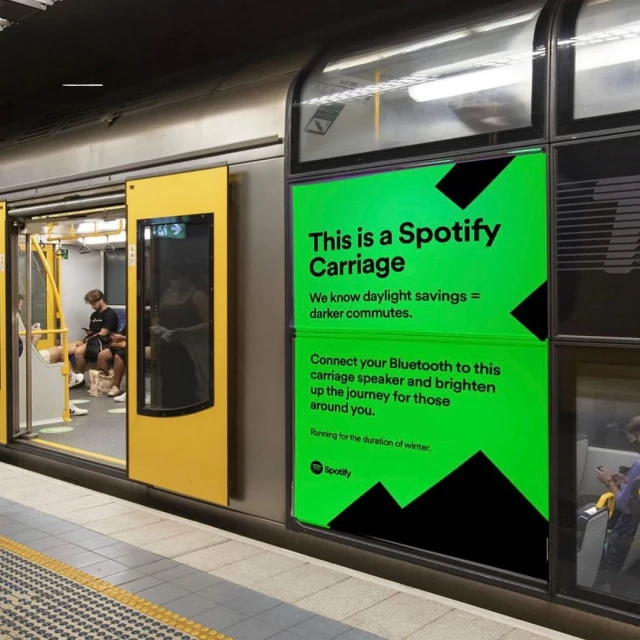 Subway car interior features a yellow door and a green Spotify ad. Two people are seated inside the car. Train interior.