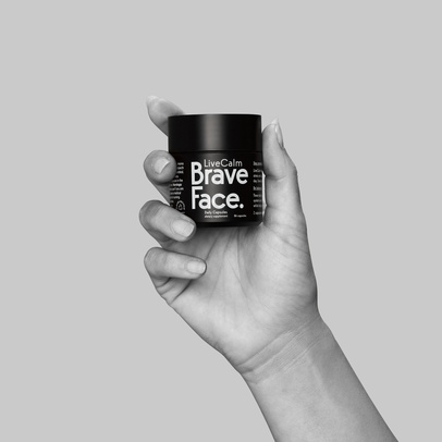 Black bottle of LiveCalm Brave Face capsules being held in a person's hand against a gray background in a studio shot.