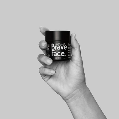 Black bottle of LiveCalm Brave Face capsules being held in a person's hand against a gray background in a studio shot.