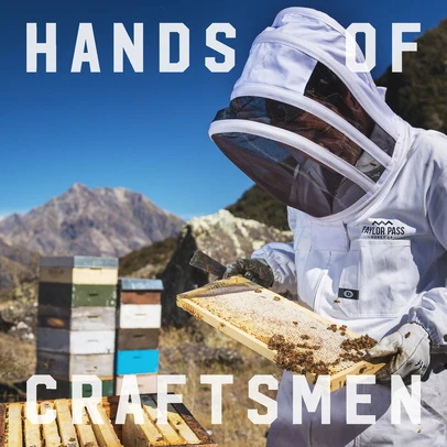 Beekeeper in protective suit inspects honeycomb in rural setting with mountains and stacked hives under blue sky. Taylor Pass Honey Co. branding.