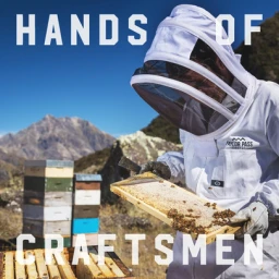 Beekeeper in a white protective suit inspecting a honeycomb frame outdoors with stacked beehives and mountains