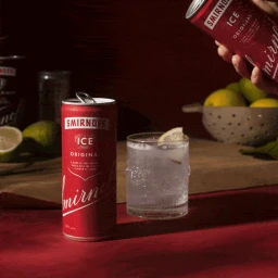 Red Smirnoff Ice can standing next to a glass of iced beverage with a lemon slice on a table with limes and a colander.