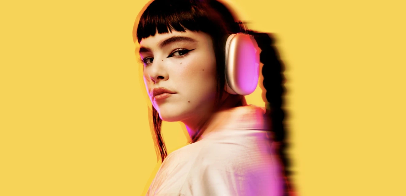 Portrait of a woman with bangs wearing wireless headphones against a yellow backdrop with pink colour effects, looking at the camera.
