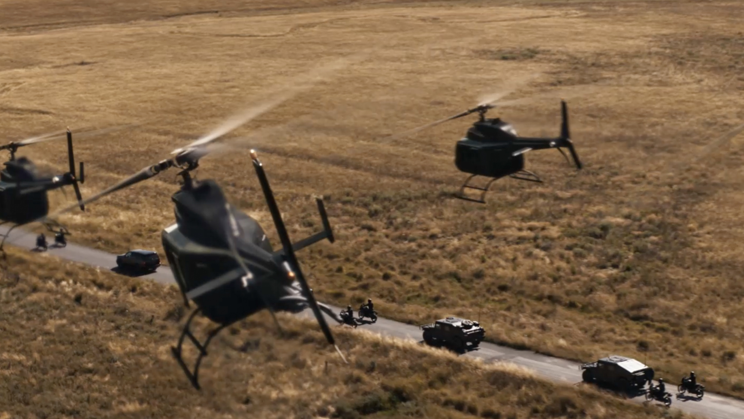 Aerial shot of helicopters flying over a rural landscape, following a convoy of vehicles on a road, creating a dramatic scene.