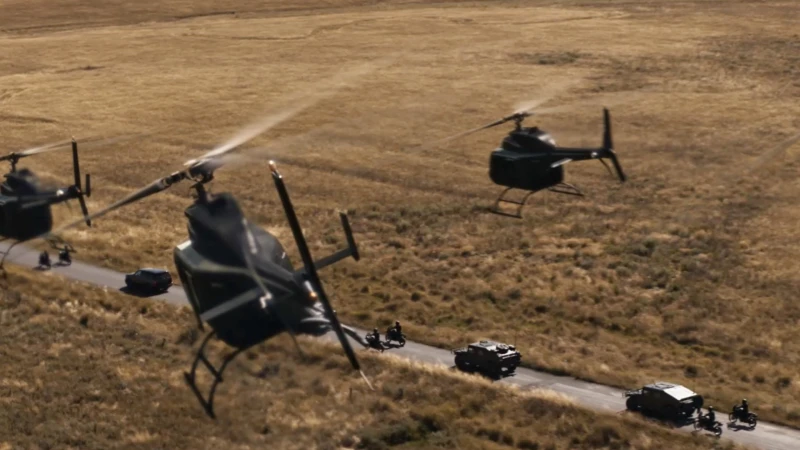Aerial shot of helicopters flying over a rural landscape, following a convoy of vehicles on a road, creating a dramatic scene.