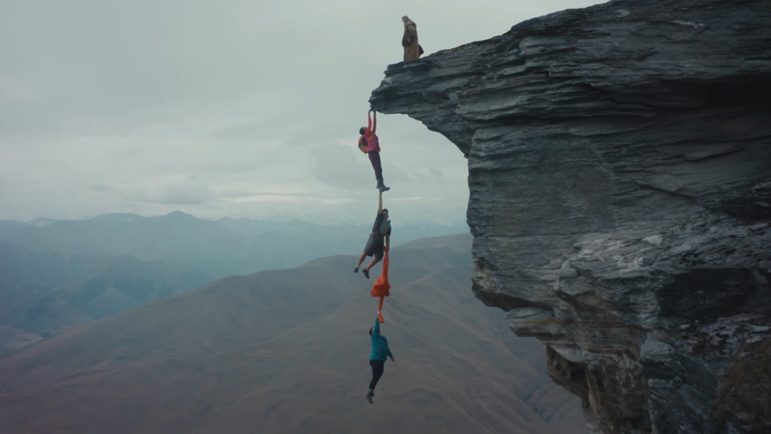 Three people hanging off a cliff face, holding onto one another, with one person on top of the cliff, overlooking a mountain range.