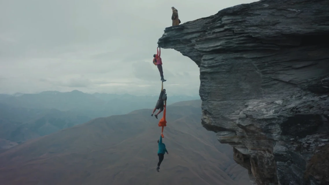Three people hanging off a cliff face, holding onto one another, with one person on top of the cliff, overlooking a mountain range.