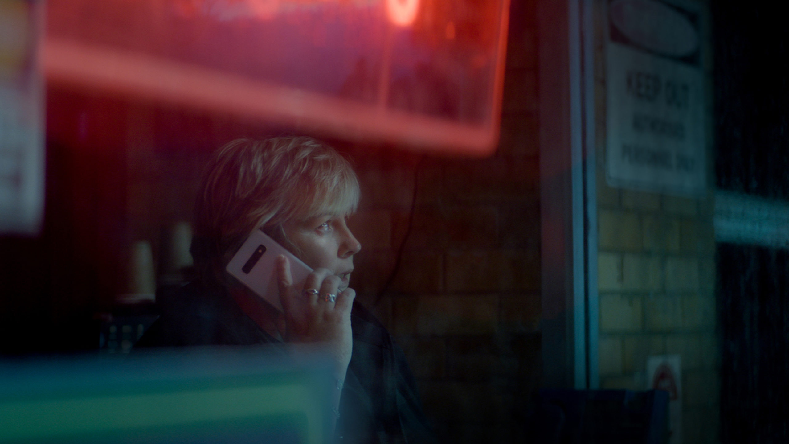 Adult woman with blonde hair is shown in a medium shot while talking on her smartphone indoors against a brick wall backdrop.