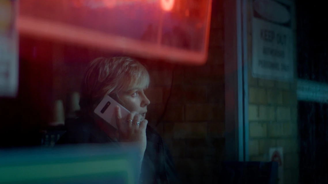 Adult woman with blonde hair is shown in a medium shot while talking on her smartphone indoors against a brick wall backdrop.