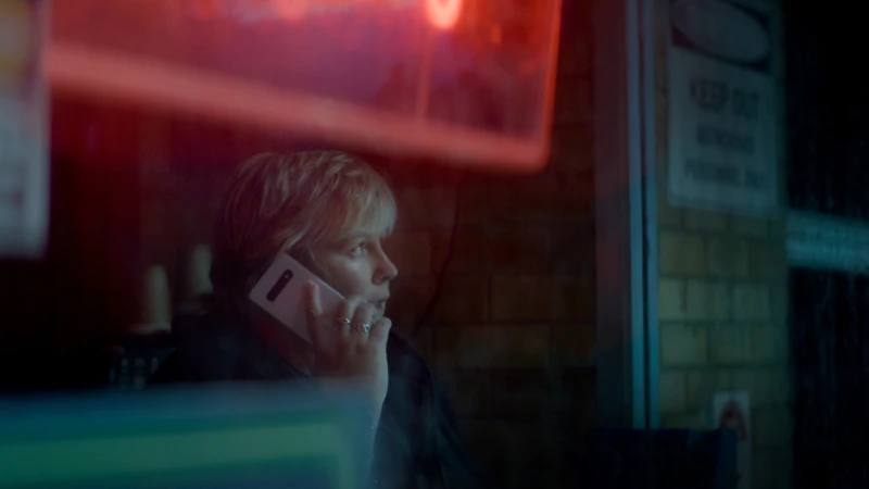 Adult woman with blonde hair is shown in a medium shot while talking on her smartphone indoors against a brick wall backdrop.