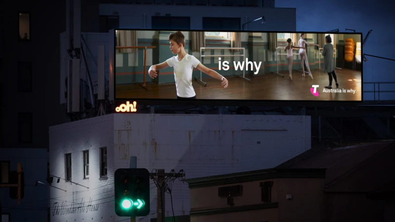 Billboard at night showing a boy ballet dancer on the left and two ballerinas with an instructor on the right; advertisement for Telstra.