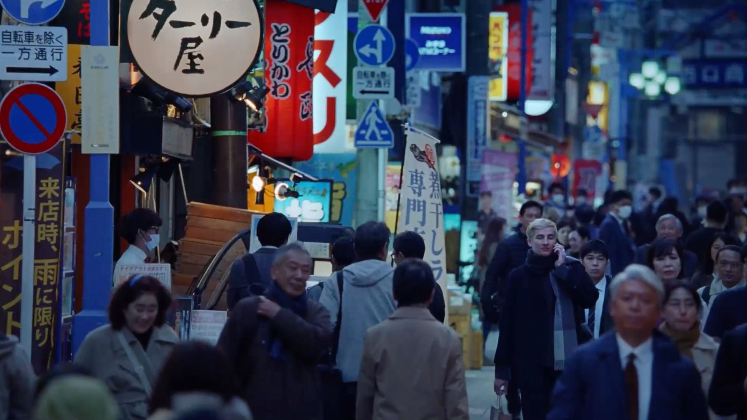 Busy Tokyo street scene with crowds of people walking by various shops and signs in the evening with a soft blue color grade.