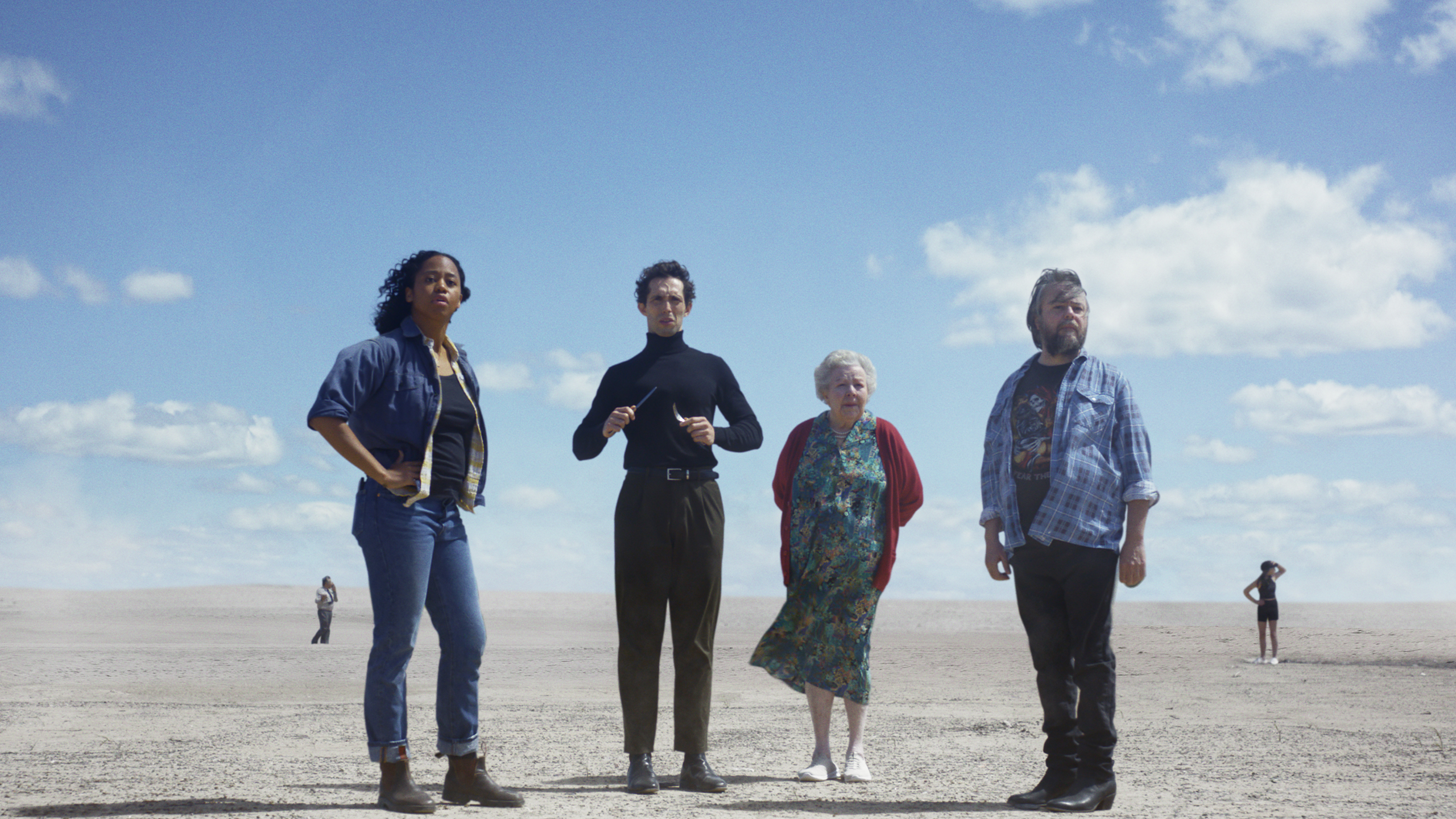 Four people stand on a desert landscape under a blue sky with clouds; a conductor holding batons is in the center.