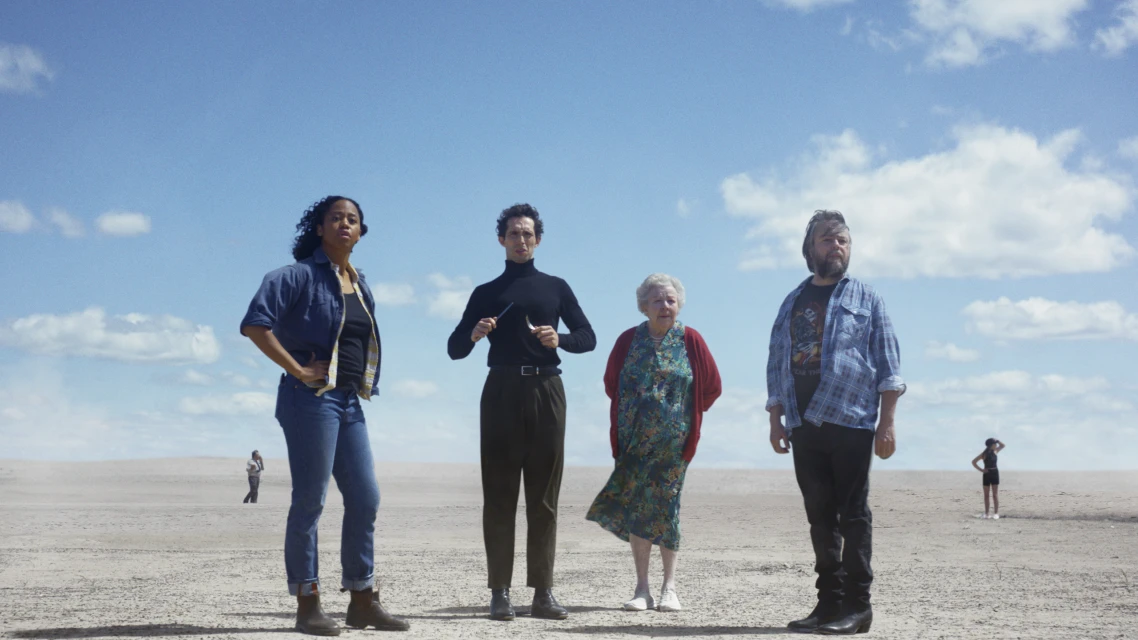 Four people stand on a desert landscape under a blue sky with clouds; a conductor holding batons is in the center.
