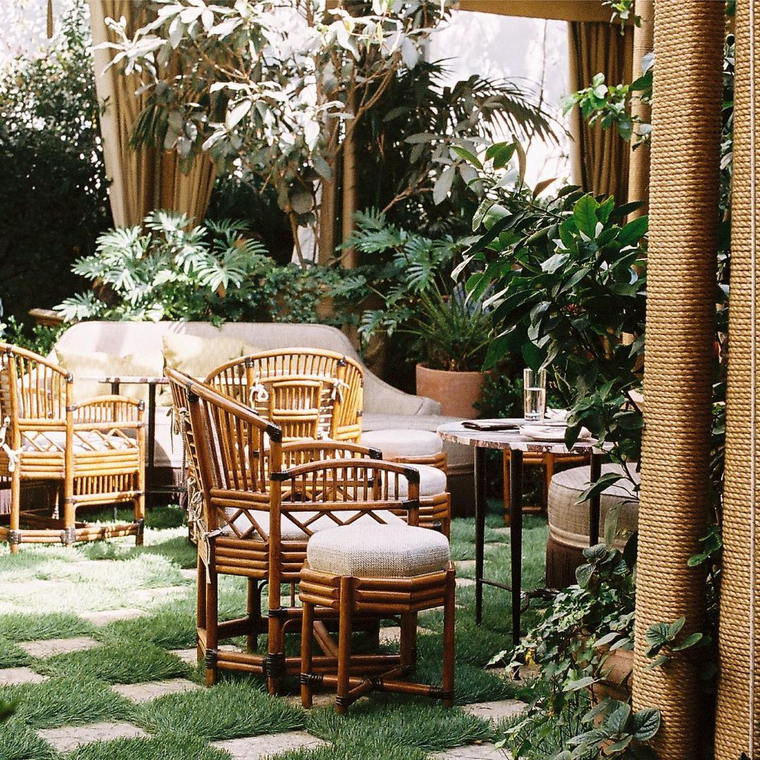 Outdoor patio area with rattan furniture set, lush green plants, grass-and-stone flooring, and soft natural lighting.