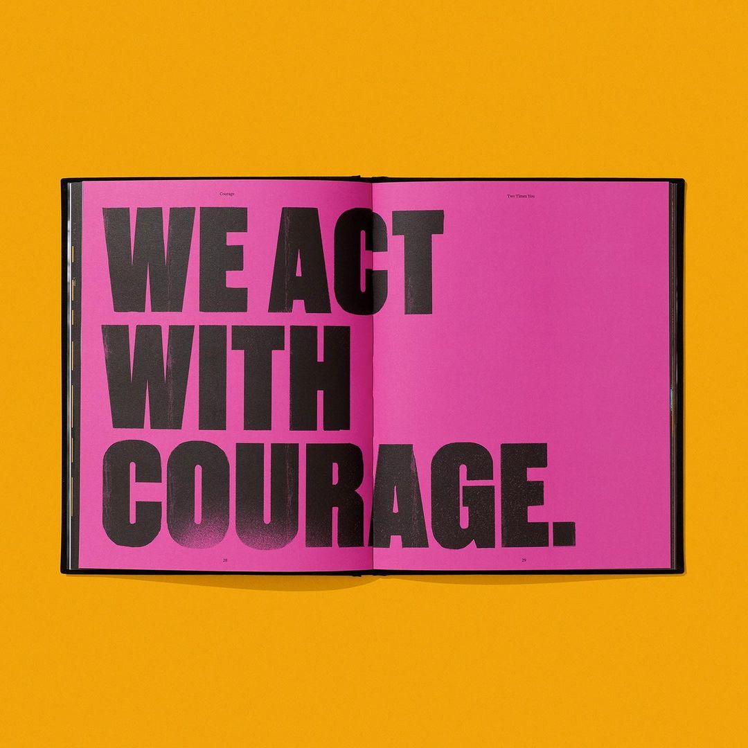Open book displaying a text-based design on pink pages against a yellow background, featuring the words 'WE ACT WITH COURAGE'.