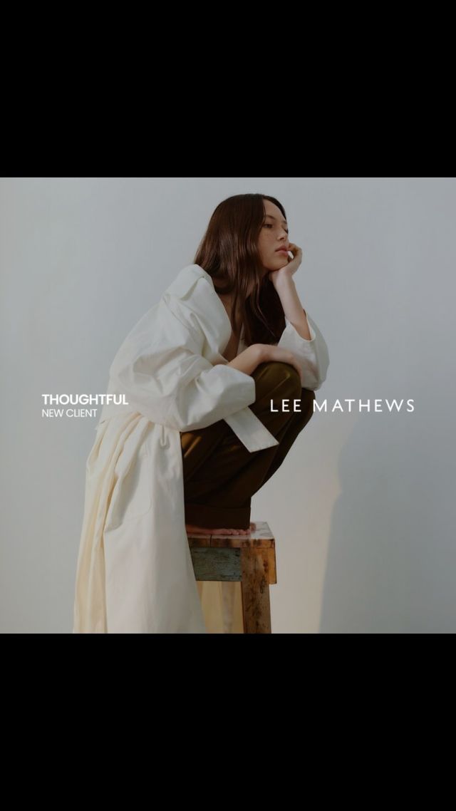 Candid studio shot of a brunette woman in a white coat crouching on a wooden stool, resting her head on her hand in thought.