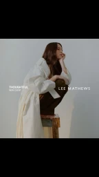 Woman in an oversized white trench coat crouching on a wooden stool against a seamless white background.