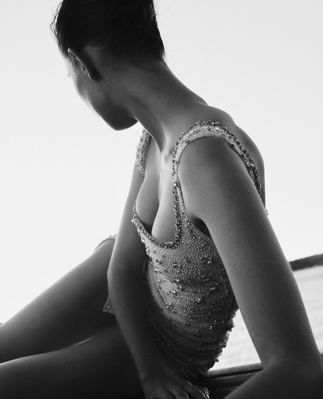 Monochrome portrait of a woman with short hair wearing an embellished garment, sitting outdoors with a bright sky in the background.