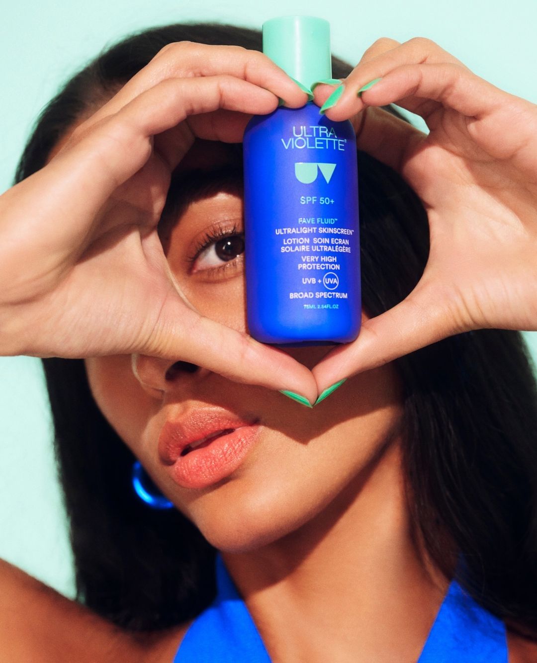 A woman holds a blue bottle of Ultra Violette sunscreen in front of her face, framing it with her fingers, against a light blue background.