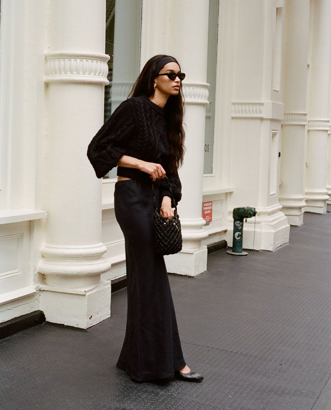 An adult female poses on a city street wearing a black headband, sunglasses, a cable knit sweater, a long skirt, and ballet flats.