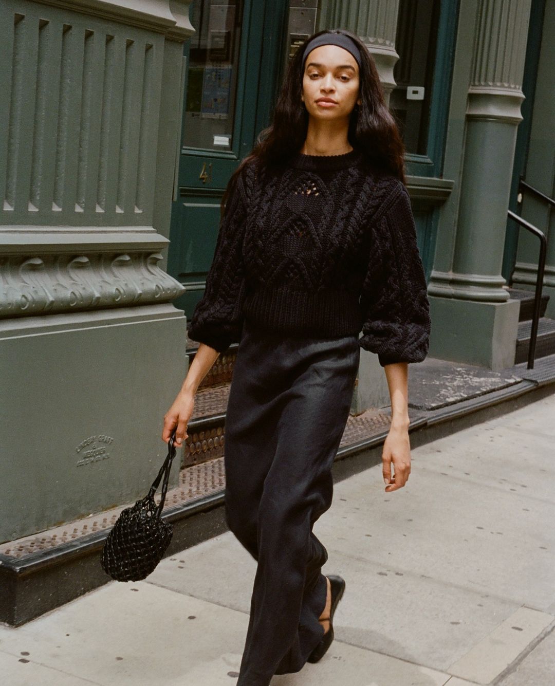 A woman in a knitted sweater and pants walks along a city sidewalk carrying a black mesh bag in an outdoor fashion shot.